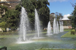 panorama da Praça da Liberdade, ponto turístico da região centro-sul de Belo Horizonte onde há uma concentração de museus e eventos culturais. Em destaque está a fonte luminosa e o coreto, ao fundo está o edifício do CCBB, Centro Cultural Banco do Brasil. Conteúdo textual: Agosto Lilás - PBH cria protocolo para proteger mulheres em espaços de lazer e turismo.