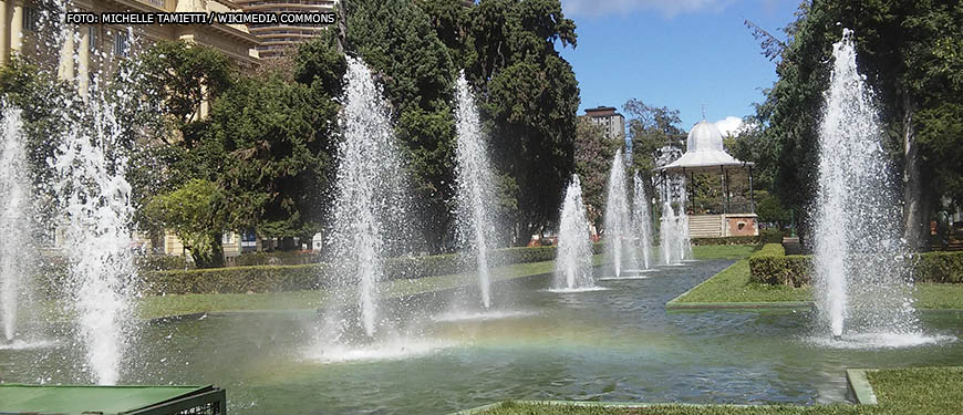 panorama da Praça da Liberdade, ponto turístico da região centro-sul de Belo Horizonte onde há uma concentração de museus e eventos culturais. Em destaque está a fonte luminosa e o coreto, ao fundo está o edifício do CCBB, Centro Cultural Banco do Brasil. Conteúdo textual: Agosto Lilás - PBH cria protocolo para proteger mulheres em espaços de lazer e turismo.