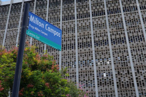 Em primeiro plano, há uma placa de rua com a identificação "Praça Milton Campos", ao fundo está a fachada de um grande edifício de concreto, com por várias aberturas retangulares em um padrão repetitivo. No canto inferior esquerdo, há uma árvore com folhas verdes e flores rosadas.