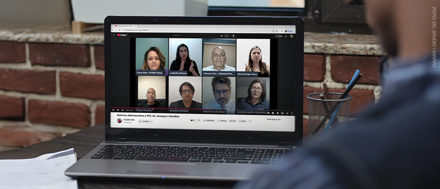 Imagem Acessível: A imagem mostra um homem sentada à mesa assistindo, em um notebook, a uma live no YouTube com oito participantes em janelas de vídeo. O título da transmissão é “Reforma Administrativa e PEC 66: ameaças e desafios”. No rodapé da imagem, em destaque, está o texto: “SINJUS denuncia riscos contidos na Reforma Administrativa e na PEC 66”.