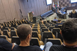 A imagem mostra um grande auditório com pessoas sentadas em cadeiras assistindo atentamente a uma reunião. As cadeiras estão dispostas em filas e à frente delas há uma grande mesa em formato de um círculo aberto, onde estão reunidos os desembargadores.