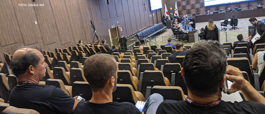 A imagem mostra um grande auditório com pessoas sentadas em cadeiras assistindo atentamente a uma reunião. As cadeiras estão dispostas em filas e à frente delas há uma grande mesa em formato de um círculo aberto, onde estão reunidos os desembargadores.