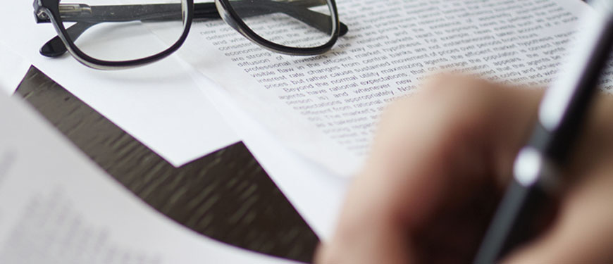 Descrição da imagem: uma mão desfocada em primeiro plano escreve em um documento. ao fundo com mais foco vemos um óculos por cima de outras folhas escritas.
