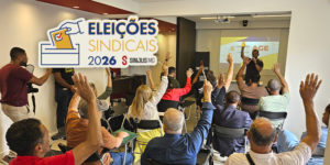 A imagem registra o momento da Assembleia-Geral Extraordinária (AGE) do SINJUS-MG, na qual foi definida a composição da Junta Eleitoral que conduzirá as Eleições Sindicais 2026. Na foto, filiadas e filiados aparecem com as mãos levantadas, votando e participando ativamente da decisão coletiva. Ao fundo, um telão exibe o tema da AGE. O texto central informa: "Definida a composição da Junta Eleitoral que irá conduzir as Eleições 2026 do SINJUS". O selo “Eleições Sindicais 2026” complementa a identidade visual da campanha, simbolizando transparência, representatividade e participação da categoria.