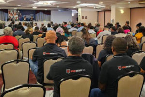 Imagem Acessível: Vista do auditório com participantes do seminário da CSPB, eles estão sentados e de costas. Conteúdo textual: Greve e negociação coletiva - SINJUS participa de seminário da CSPB em Brasília sobre Convenção 151 da OIT.