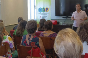 Foto vespertina da reunião do NAP, se vê uma sala com cadeiras, nelas estão sentados os aposentados e pensionistas que integram o núcleo, à frente deles se vê o palestrante.