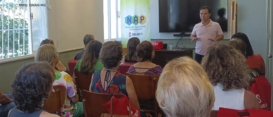 Foto vespertina da reunião do NAP, se vê uma sala com cadeiras, nelas estão sentados os aposentados e pensionistas que integram o núcleo, à frente deles se vê o palestrante.