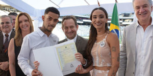 : Montagem fotográfica de um casamento coletivo realizado em um estádio de futebol (Arena MRV). Na parte superior, estão o presidente do TJMG, Desembargador Luiz Carlos de Azevedo Corrêa Jr. e Sérgio Coelho, presidente do Clube Atlético Mineiro acompanhados por um casal sorridente que exibe a certidão de casamento. Na parte inferior, várias cenas mostram dezenas de casais vestidos majoritariamente de branco trocando beijos e abraços durante a cerimônia, criando um clima de celebração coletiva.