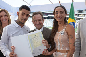 : Montagem fotográfica de um casamento coletivo realizado em um estádio de futebol (Arena MRV). Na parte superior, estão o presidente do TJMG, Desembargador Luiz Carlos de Azevedo Corrêa Jr. e Sérgio Coelho, presidente do Clube Atlético Mineiro acompanhados por um casal sorridente que exibe a certidão de casamento. Na parte inferior, várias cenas mostram dezenas de casais vestidos majoritariamente de branco trocando beijos e abraços durante a cerimônia, criando um clima de celebração coletiva.