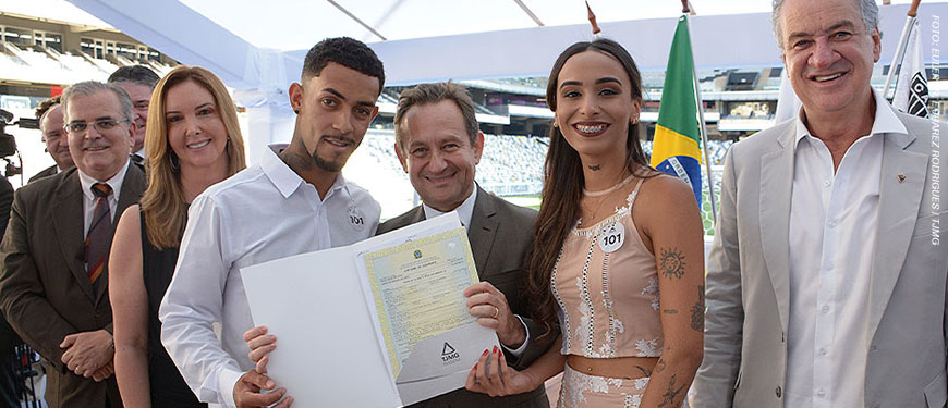 : Montagem fotográfica de um casamento coletivo realizado em um estádio de futebol (Arena MRV). Na parte superior, estão o presidente do TJMG, Desembargador Luiz Carlos de Azevedo Corrêa Jr. e Sérgio Coelho, presidente do Clube Atlético Mineiro acompanhados por um casal sorridente que exibe a certidão de casamento. Na parte inferior, várias cenas mostram dezenas de casais vestidos majoritariamente de branco trocando beijos e abraços durante a cerimônia, criando um clima de celebração coletiva.