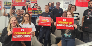 A fotografia mostra um grupo de servidores e servidoras reunidos, segurando cartazes em apoio à valorização do Auxílio-Saúde. As mensagens nos cartazes incluem frases como “Auxílio-Saúde digno já!”, “Isonomia no Auxílio-Saúde” e “A saúde do servidor também importa”. Ao fundo, há uma faixa com o logotipo do SINJUS-MG. O clima é de mobilização e cobrança por justiça na concessão do benefício.