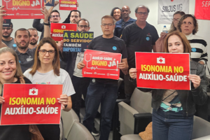 A fotografia mostra um grupo de servidores e servidoras reunidos, segurando cartazes em apoio à valorização do Auxílio-Saúde. As mensagens nos cartazes incluem frases como “Auxílio-Saúde digno já!”, “Isonomia no Auxílio-Saúde” e “A saúde do servidor também importa”. Ao fundo, há uma faixa com o logotipo do SINJUS-MG. O clima é de mobilização e cobrança por justiça na concessão do benefício.