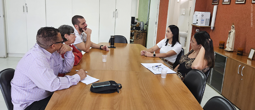 A imagem mostra uma sala de reuniões simples, onde cinco pessoas estão sentadas ao redor de uma mesa de madeira, conversando durante uma visita técnica ao Centro Odontológico do Ipsemg, em Belo Horizonte. À direita, duas gestoras da unidade ouvem representantes do Conselho de Beneficiários do Ipsemg (CBI), entre eles o coordenador-geral do SINJUS-MG, Alexandre Pires.