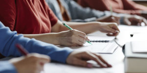 A imagem mostra uma fileira de estudantes sentados em sala de aula, escrevendo em cadernos e fazendo anotações. O foco está nas mãos de uma pessoa escrevendo com caneta. Sobre a imagem, o texto diz: “EJEF e PUC Minas firmam parceria para implantação de mestrado e doutorado no TJMG”. No canto superior esquerdo, está o título “Oportunidade” e, no canto inferior, o logotipo do SINJUS-MG.