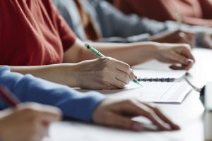 A imagem mostra uma fileira de estudantes sentados em sala de aula, escrevendo em cadernos e fazendo anotações. O foco está nas mãos de uma pessoa escrevendo com caneta. Sobre a imagem, o texto diz: “EJEF e PUC Minas firmam parceria para implantação de mestrado e doutorado no TJMG”. No canto superior esquerdo, está o título “Oportunidade” e, no canto inferior, o logotipo do SINJUS-MG.