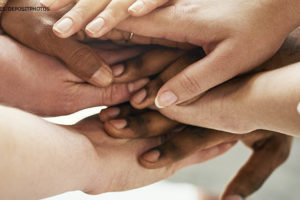 Imagem Acessível: Mãos de pessoas de diversas etnias estão posicionadas umas sobre as outras, em um gesto de união e apoio.