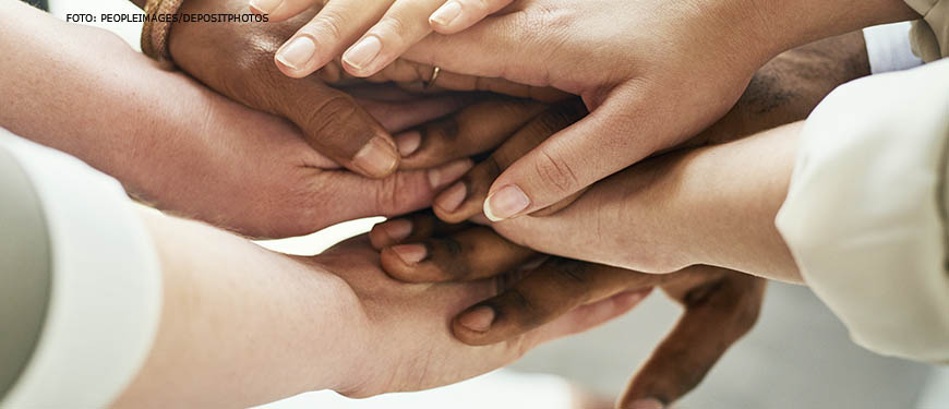 Imagem Acessível: Mãos de pessoas de diversas etnias estão posicionadas umas sobre as outras, em um gesto de união e apoio.