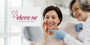 Imagem acessível: A Imagem divulga o novo convênio do SINJUS-MG, mostrando ao fundo a foto de uma clínica de estética focando em uma mulher adulta sorrindo enquanto se olha em um espelho que segura, aparentando satisfação com a própria aparência, e ao lado dela, uma profissional mais velha toca suavemente seu rosto com expressão acolhedora, em um ambiente claro que transmite cuidado e bem-estar. No centro da imagem, está o logotipo do convênio “Eleve-se, Estética e Bem-Estar”, com símbolo de borboleta, e logo abaixo lê-se o texto em destaque: “Fique em dia com a sua autoestima com o novo convênio do SINJUS”, enquanto no topo aparece o texto “Clínica de Estética”.