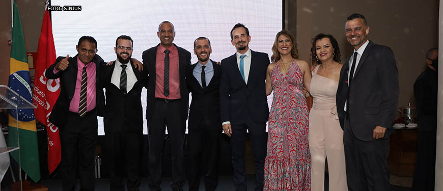 Grupo de 2 mulheres e 6 homens em trajes sociais (diretores do SINJUS) reunidos em um palco com uma tela de led ao fundo.