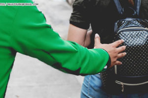 Pessoa encapuzada se aproxima pelas costas de uma mulher com mochila, como se fosse roubá-la.