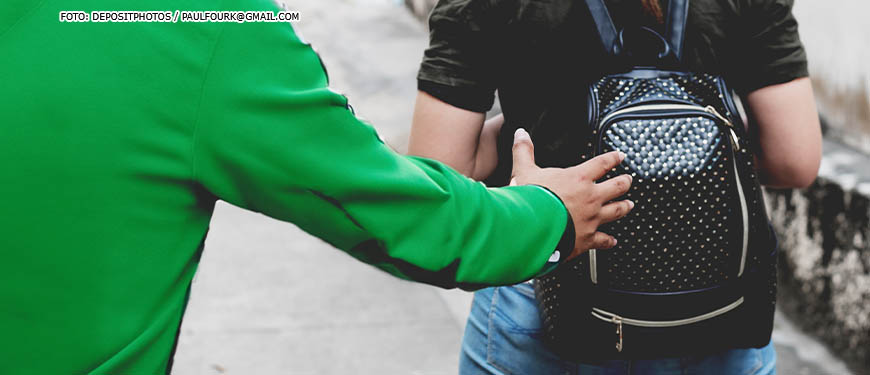 Pessoa encapuzada se aproxima pelas costas de uma mulher com mochila, como se fosse roubá-la.