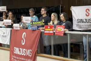 Imagem Acessível: A imagem mostra uma galeria do Plenário da ALMG com servidores e representantes sindicais reunidos em manifestação. Eles seguram cartazes com dizeres como “Auxílio-Saúde Digno Já”, “Isonomia no Auxílio-Saúde” e “A saúde do servidor também é importante”, em apoio à aprovação do PL do Auxílio-Saúde. Três faixas de sindicatos estão afixadas na mureta da galeria. O clima é de mobilização e conquista após a aprovação do projeto em 1º turno.