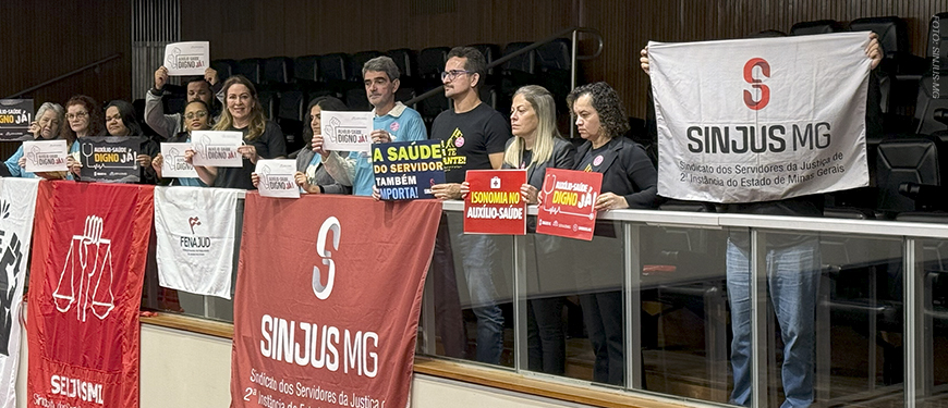 Imagem Acessível: A imagem mostra uma galeria do Plenário da ALMG com servidores e representantes sindicais reunidos em manifestação. Eles seguram cartazes com dizeres como “Auxílio-Saúde Digno Já”, “Isonomia no Auxílio-Saúde” e “A saúde do servidor também é importante”, em apoio à aprovação do PL do Auxílio-Saúde. Três faixas de sindicatos estão afixadas na mureta da galeria. O clima é de mobilização e conquista após a aprovação do projeto em 1º turno.
