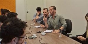 Reunião com cerca de dez pessoas sentadas ao redor de uma mesa, em ambiente interno e iluminado. No centro da imagem, há dois dirigentes do SINJUS-MG: o coordenador-geral, Alexandre Pires e o diretor de Assuntos Jurídicos, Felipe Rodrigues. Ambos falam com expressividade e gesticulam enquanto seguram papéis. Os demais participantes observam atentamente ou fazem anotações.