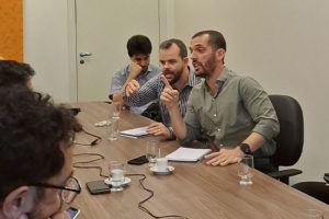 Reunião com cerca de dez pessoas sentadas ao redor de uma mesa, em ambiente interno e iluminado. No centro da imagem, há dois dirigentes do SINJUS-MG: o coordenador-geral, Alexandre Pires e o diretor de Assuntos Jurídicos, Felipe Rodrigues. Ambos falam com expressividade e gesticulam enquanto seguram papéis. Os demais participantes observam atentamente ou fazem anotações.