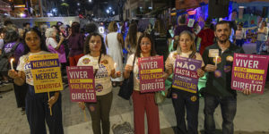 Imagem acessível: Fotografia de ato noturno realizado na Rua Sapucaí, em Belo Horizonte, pelos 20 anos da Lei Maria da Penha. Em primeiro plano, cinco participantes seguram velas e cartazes com mensagens como “Viver sem violência é um direito” e “Nenhuma mulher merece virar uma estatística”. Entre eles, estão dirigentes do SINJUS-MG. Ao fundo, outras pessoas participam do ato, em clima de manifestação.