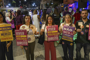 Imagem acessível: Fotografia de ato noturno realizado na Rua Sapucaí, em Belo Horizonte, pelos 20 anos da Lei Maria da Penha. Em primeiro plano, cinco participantes seguram velas e cartazes com mensagens como “Viver sem violência é um direito” e “Nenhuma mulher merece virar uma estatística”. Entre eles, estão dirigentes do SINJUS-MG. Ao fundo, outras pessoas participam do ato, em clima de manifestação.