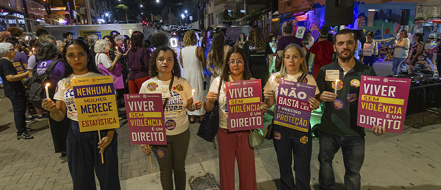 Imagem acessível: Fotografia de ato noturno realizado na Rua Sapucaí, em Belo Horizonte, pelos 20 anos da Lei Maria da Penha. Em primeiro plano, cinco participantes seguram velas e cartazes com mensagens como “Viver sem violência é um direito” e “Nenhuma mulher merece virar uma estatística”. Entre eles, estão dirigentes do SINJUS-MG. Ao fundo, outras pessoas participam do ato, em clima de manifestação.