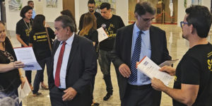 A imagem mostra um grupo de pessoas usando camisetas pretas com a identidade do SINJUS-MG entregando cartas a representantes de terno no saguão do TJMG. O texto na imagem diz: “Comissão de Excedentes entrega carta cobrando mais vagas na PV 2023 durante sessão do Órgão Especial”.