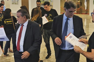 A imagem mostra um grupo de pessoas usando camisetas pretas com a identidade do SINJUS-MG entregando cartas a representantes de terno no saguão do TJMG. O texto na imagem diz: “Comissão de Excedentes entrega carta cobrando mais vagas na PV 2023 durante sessão do Órgão Especial”.