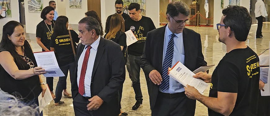 A imagem mostra um grupo de pessoas usando camisetas pretas com a identidade do SINJUS-MG entregando cartas a representantes de terno no saguão do TJMG. O texto na imagem diz: “Comissão de Excedentes entrega carta cobrando mais vagas na PV 2023 durante sessão do Órgão Especial”.