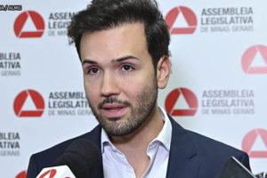 Ao fundo há um painel com o logo da ALMG disposto como um padrão de xadrez, à frente dele está um homem branco, o deputado Tadeu Leite, ele veste um paletó azul escuro com camisa social azul clara, tem barba curta e cabelo liso escovado para o alto. O homem está falando para microfones e smartphones com os logos de veículos de comunicação de Minas Gerais.