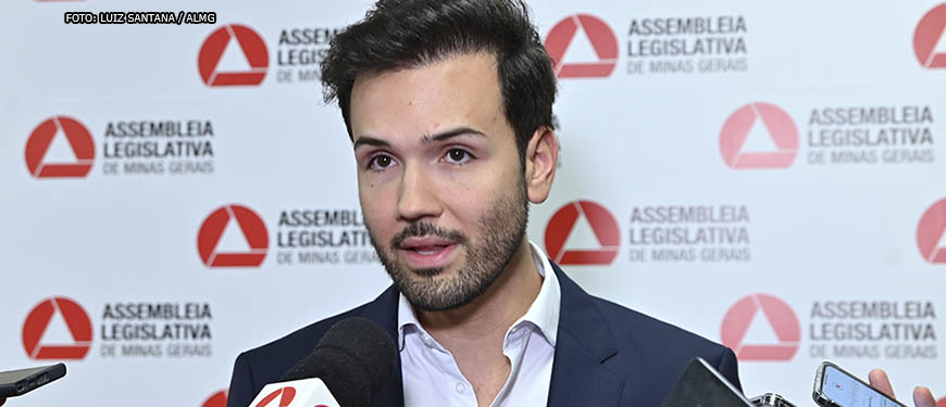 Ao fundo há um painel com o logo da ALMG disposto como um padrão de xadrez, à frente dele está um homem branco, o deputado Tadeu Leite, ele veste um paletó azul escuro com camisa social azul clara, tem barba curta e cabelo liso escovado para o alto. O homem está falando para microfones e smartphones com os logos de veículos de comunicação de Minas Gerais.