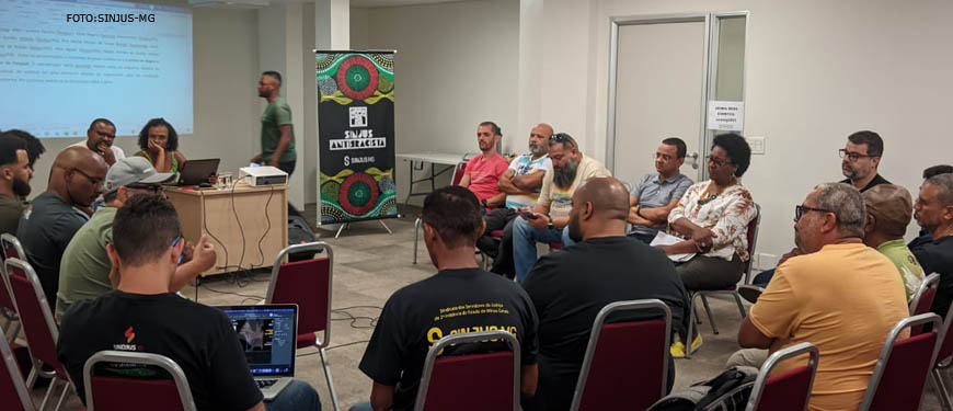 Imagem Acessível: A fotografia retrata um grupo de pessoas em uma sala em prol do Coletivo de Negras e Negros da Fenajud. Conteúdo Textual: ANTIRRACISMO COLETIVO DE NEGRAS E NEGROS DA FENAJUD REALIZA PRIMEIRO ENCONTRO EM BH