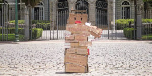 Fotografia diurna de obra feita em papelão representando uma figura feminina, com várias palavras e frases escritas nas caixas que a compõem. Ao fundo da obra, está o Palácio da Liberdade e algumas árvores.