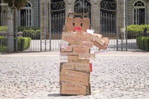 Fotografia diurna de obra feita em papelão representando uma figura feminina, com várias palavras e frases escritas nas caixas que a compõem. Ao fundo da obra, está o Palácio da Liberdade e algumas árvores.