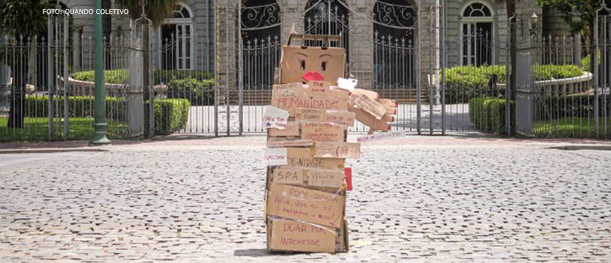 Fotografia diurna de obra feita em papelão representando uma figura feminina, com várias palavras e frases escritas nas caixas que a compõem. Ao fundo da obra, está o Palácio da Liberdade e algumas árvores.