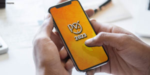 Se vê a imagem de uma pessoa segurando um celular, onde o fundo de tela do mesmo está escrito "PV 2023" na cor cinza com o fundo amarelo.