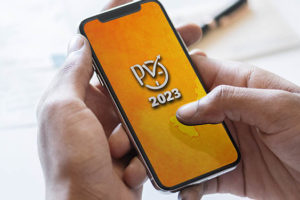 Se vê a imagem de uma pessoa segurando um celular, onde o fundo de tela do mesmo está escrito "PV 2023" na cor cinza com o fundo amarelo.