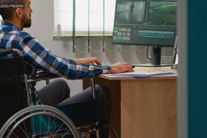 Um homem branco deficiente em cadeira de rodas trabalha em um computador. Conteúdo textual: DIREITOS - Reserva de cargos para pessoa com deficiência pode subir.