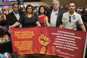 A imagem mostra um grupo de cinco pessoas segurando uma faixa vermelha em um auditório lotado, durante o seminário na Câmara dos Deputados. A faixa tem mensagens de apoio ao serviço público e ao Regime Jurídico Único, destacando a importância da estabilidade para garantir continuidade, imparcialidade e impessoalidade no serviço público. O evento discute o possível fim desse regime, e várias pessoas no auditório estão acompanhando a discussão. A Fenajud, entidade que representa servidores do Judiciário, está envolvida na mobilização.