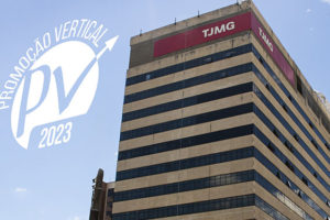 Fotografia de um grande prédio com fachada bege e janelas espelhadas escuras. No topo do edifício, há placas vermelhas com a sigla "TJMG" (Tribunal de Justiça de Minas Gerais). O céu azul com algumas nuvens aparece ao fundo. Na parte superior da imagem, há um logotipo branco com a inscrição "Promoção Vertical PV 2023" e uma seta apontando para cima. Conteúdo textual: CARREIRA - TJMG divulga decisão acerca dos pedidos de reconsideração da PV 2023.