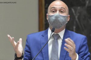 Foto do Deputado estadual Gustavo Valadares (homem de pele clara, calvo, vestindo um terno azul e usando máscara) falando ao microfone em um púlpito.