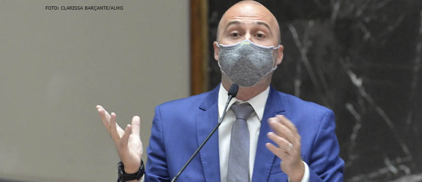 Foto do Deputado estadual Gustavo Valadares (homem de pele clara, calvo, vestindo um terno azul e usando máscara) falando ao microfone em um púlpito.
