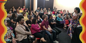 Visão do auditório lotado durante a palestra da Dra. Lívia Sant'Anna Vaz, que fala ao microfone. Ela é uma mulher negra de cabelos cacheados de comprimento médio, ela veste um vestido com estampa colorida.