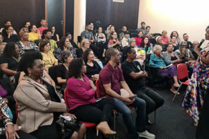 Visão do auditório lotado durante a palestra da Dra. Lívia Sant'Anna Vaz, que fala ao microfone. Ela é uma mulher negra de cabelos cacheados de comprimento médio, ela veste um vestido com estampa colorida.